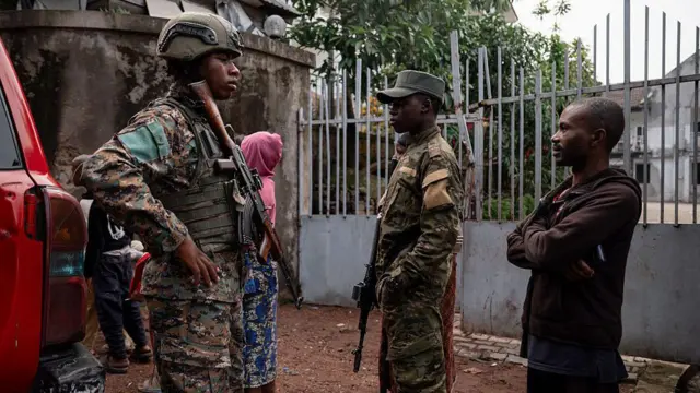 Deux militaires en uniforme, armés, accompagnés d'un civil en sweat-shirt marron et capuche, avec une femme en rose en arrière-plan
